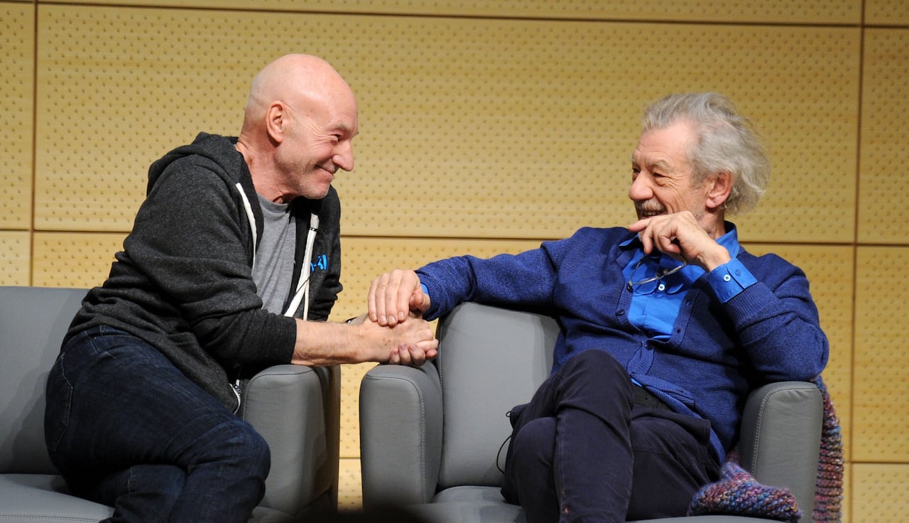 Two older men sit beside each other in armchairs. One is leaning towards the other over the arm, smiling, while the other sits back in his chair and pats the other's hand.
