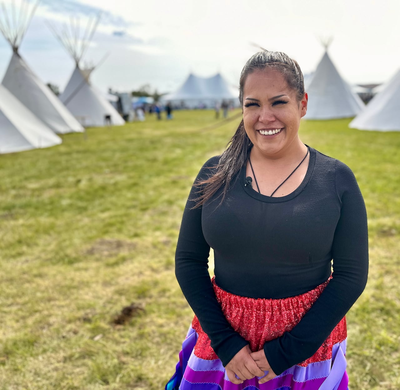 A young smiling woman in a colourful ribbon skirt smiles for the camera in front of white tipi's