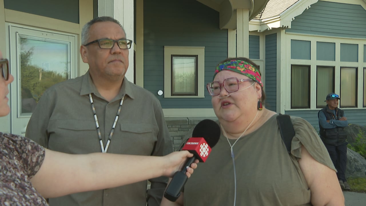 A man and woman stand shoulder-to-shoulder outside of a one-storey building.