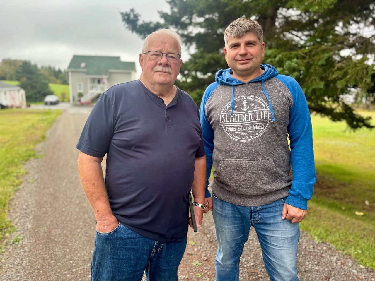 Two men stand on a laneway in front of a large tree and a house.