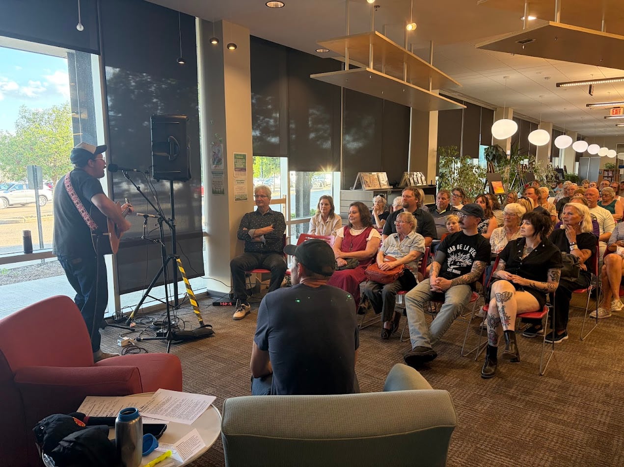 a couple dozen people sit in a room on chairs in rows as a man up front plays a guitar
