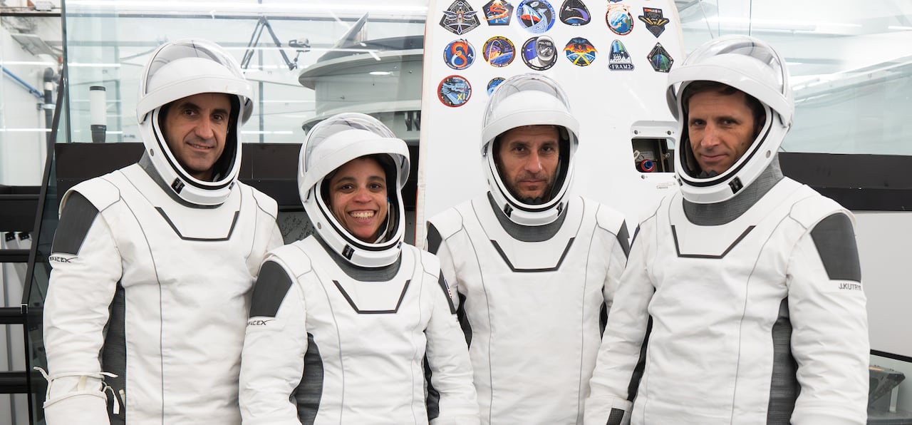 Four people in white spacesuits stand, smiling at the camera.