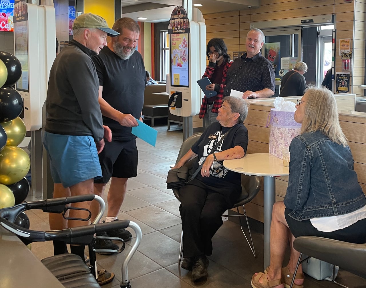 Two women sit at a table in a restaurant while people gather behind them with greeting cards in their hands.