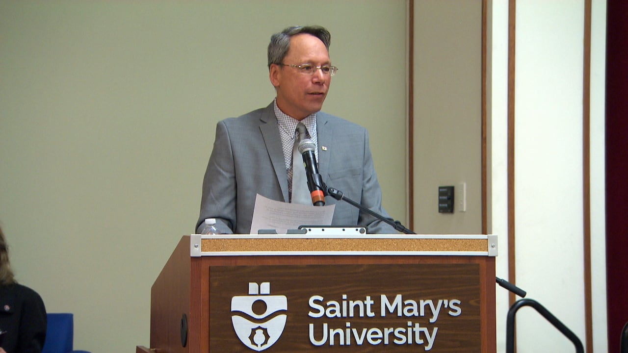 A man wearing a suit and tie is shown speaking at a podium.