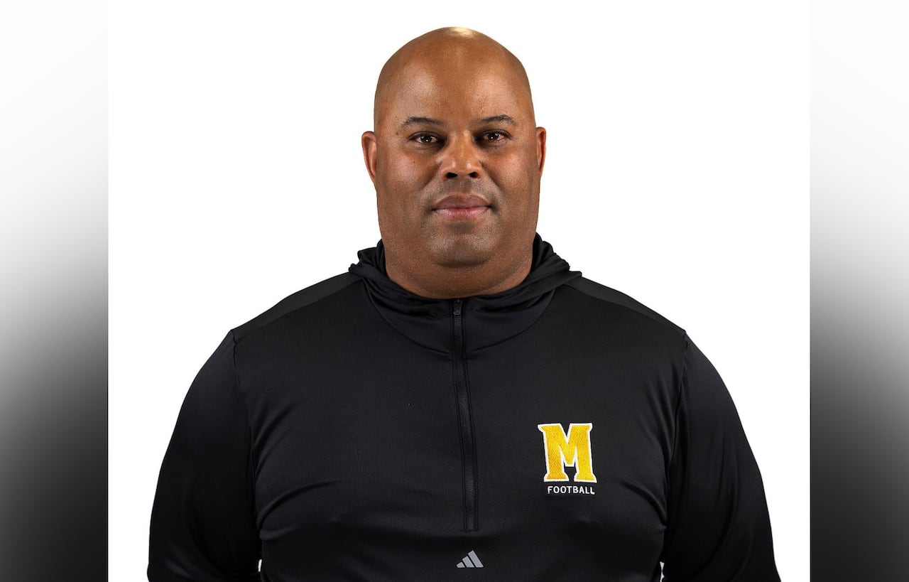 A bald man in a black hooded sweatshirt emblazoned with the letter M, is seen from the chest up.