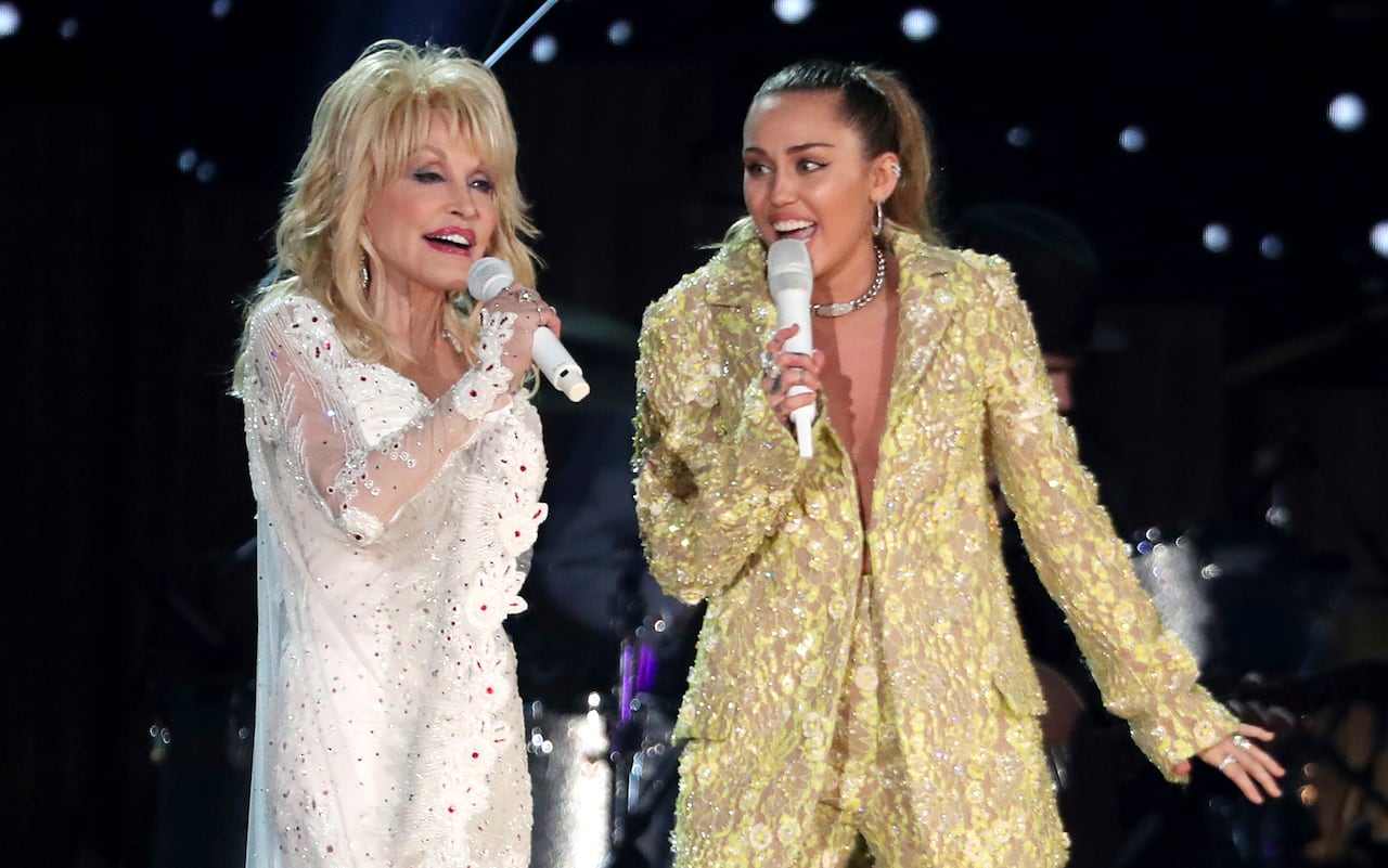 An older woman in a shiny white outfit sings onstage with a younger woman in a shiny gold outfit.