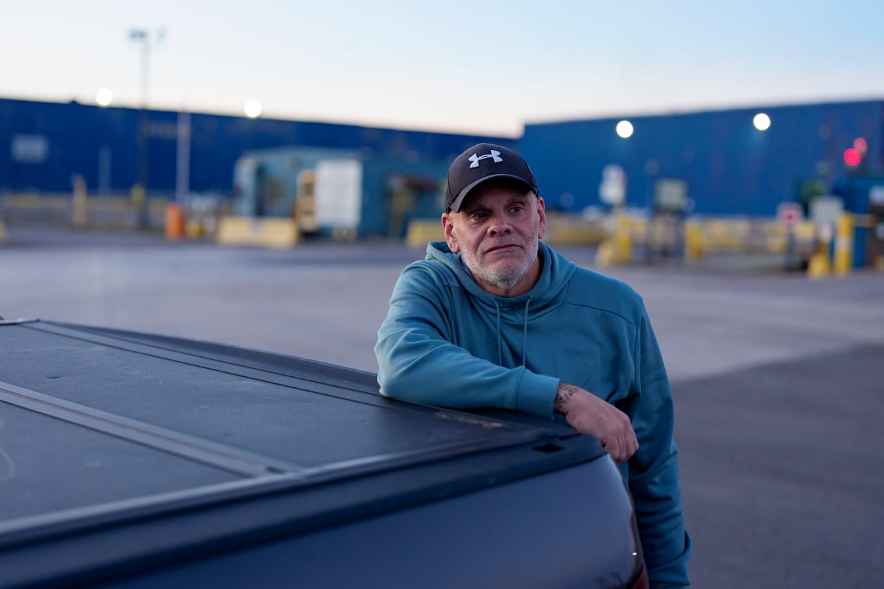 A man standing by the back of a pickup, with a blue warehouse in the distance.