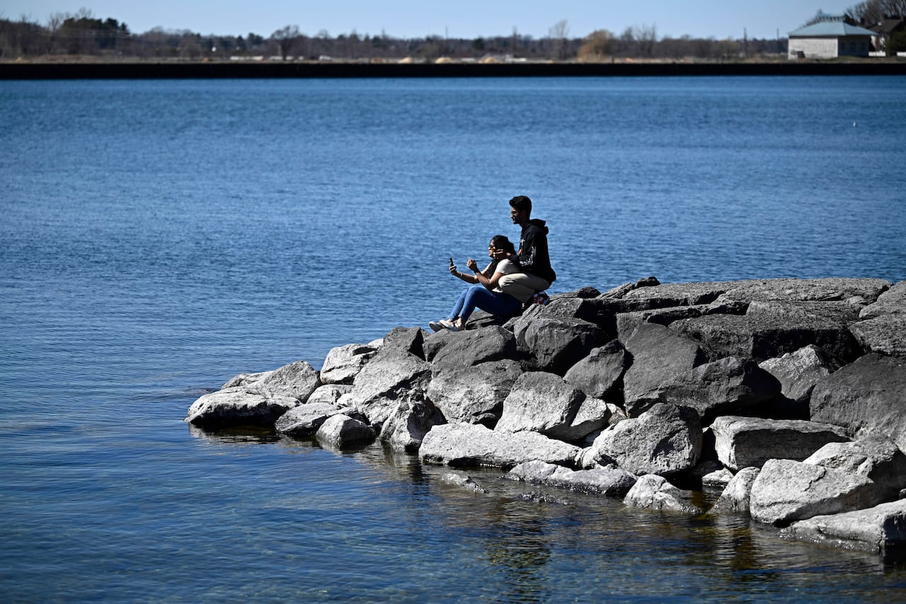 Two people sit on rocks on the shore of a Great Lake in early spring. One is taking a picture.