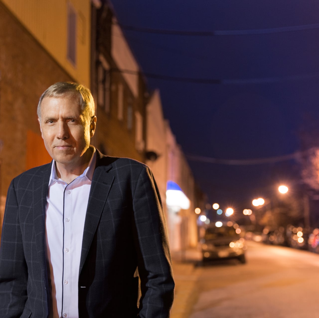 Man in suit looks in camera in front of a street lit by street lamps in the evening