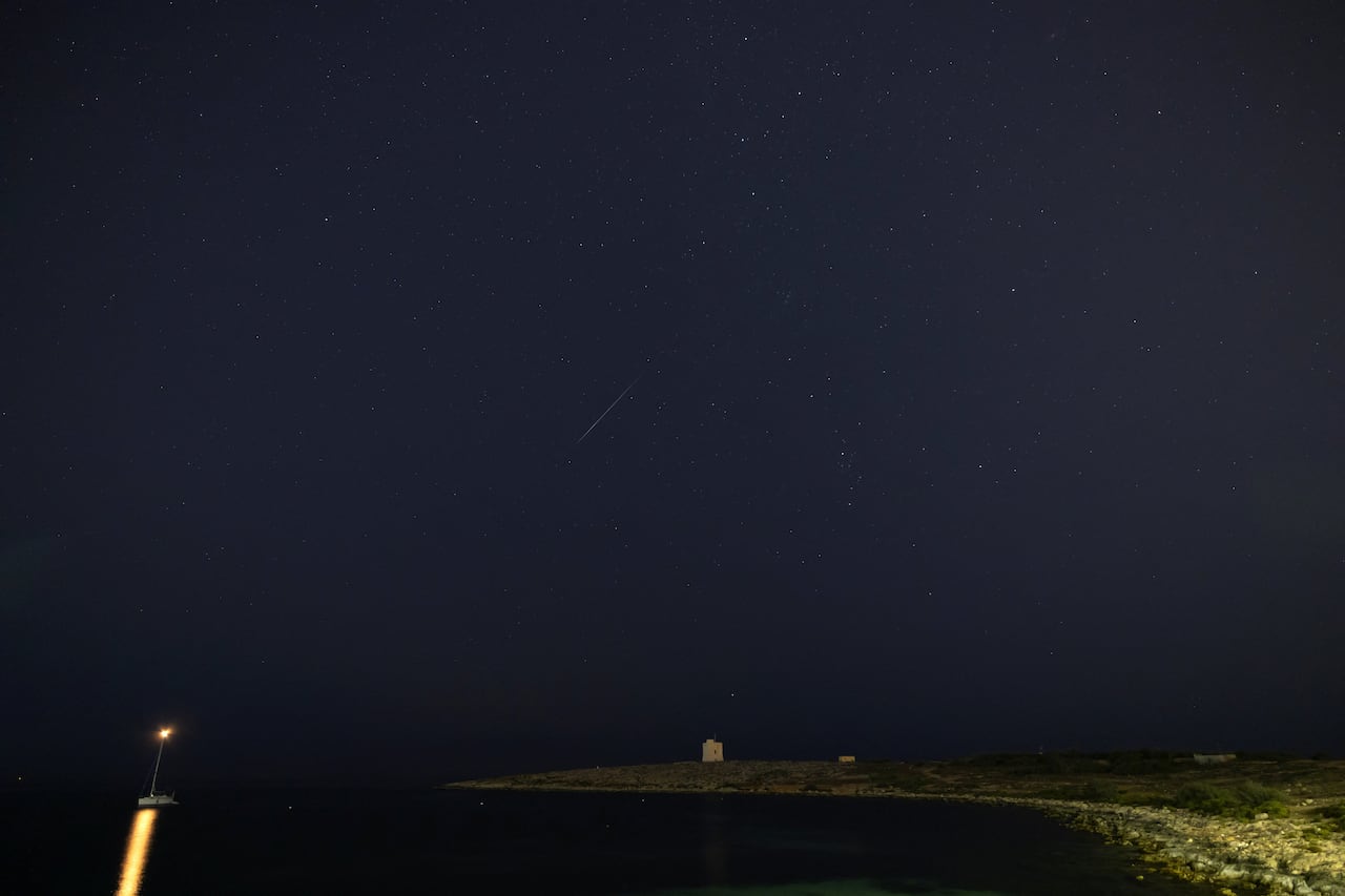 A sailboat near a shoreline under a night sky showing meteor streaks and stars.