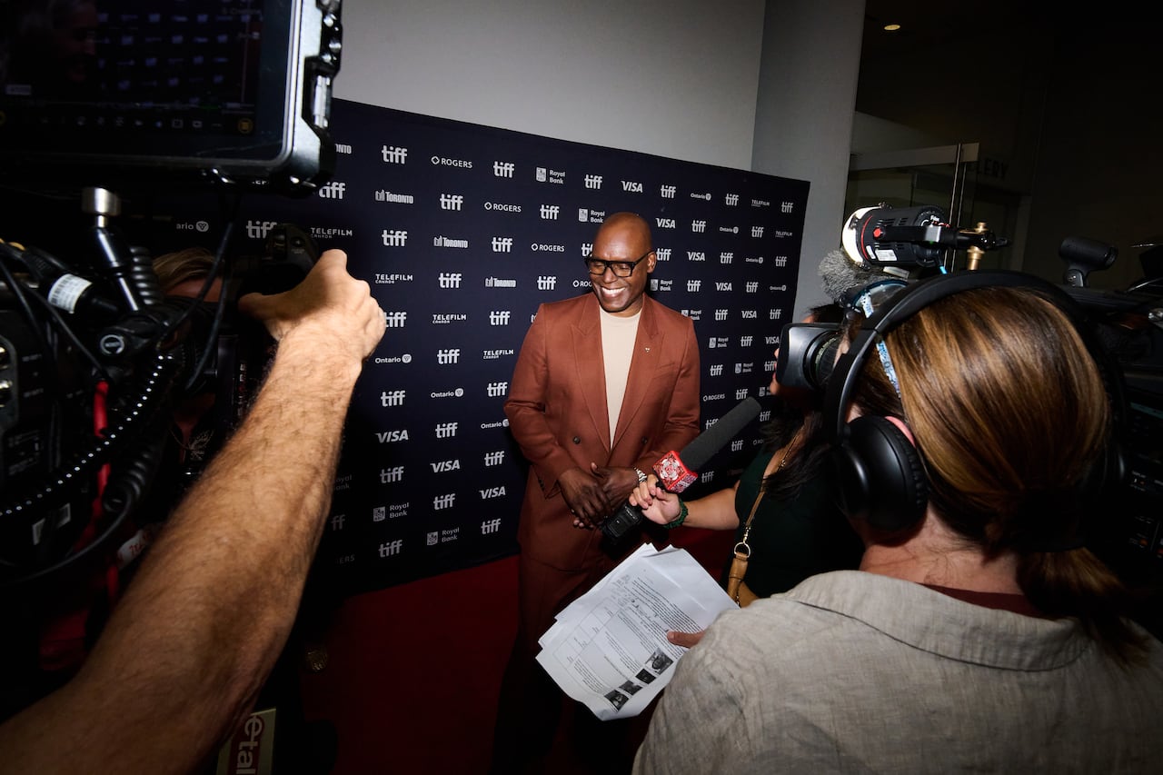 A man wearing a brown suit and glasses stands on the TIFF red carpet with cameras around him, with a black screen background behind him bearing the words "TIFF."