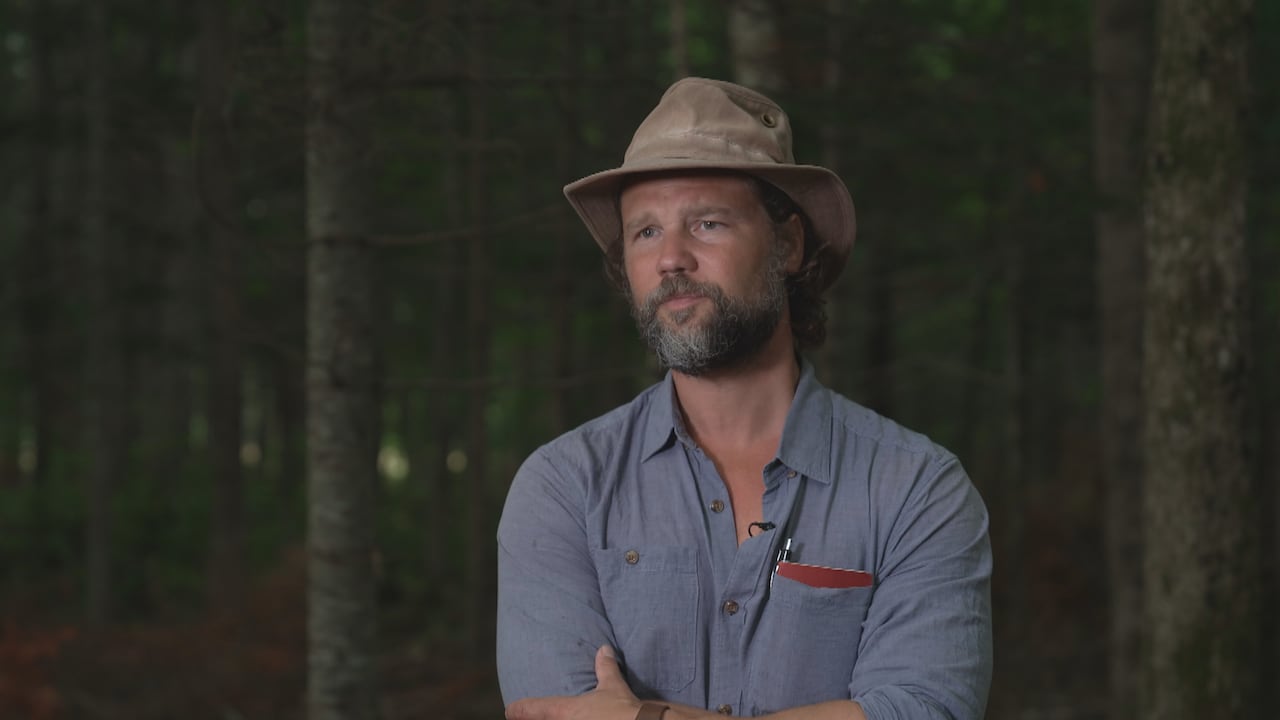 A white man with a beard, wearing a Tilley hat and a blue collared shirt, standing in front of a background of evergreen trees.