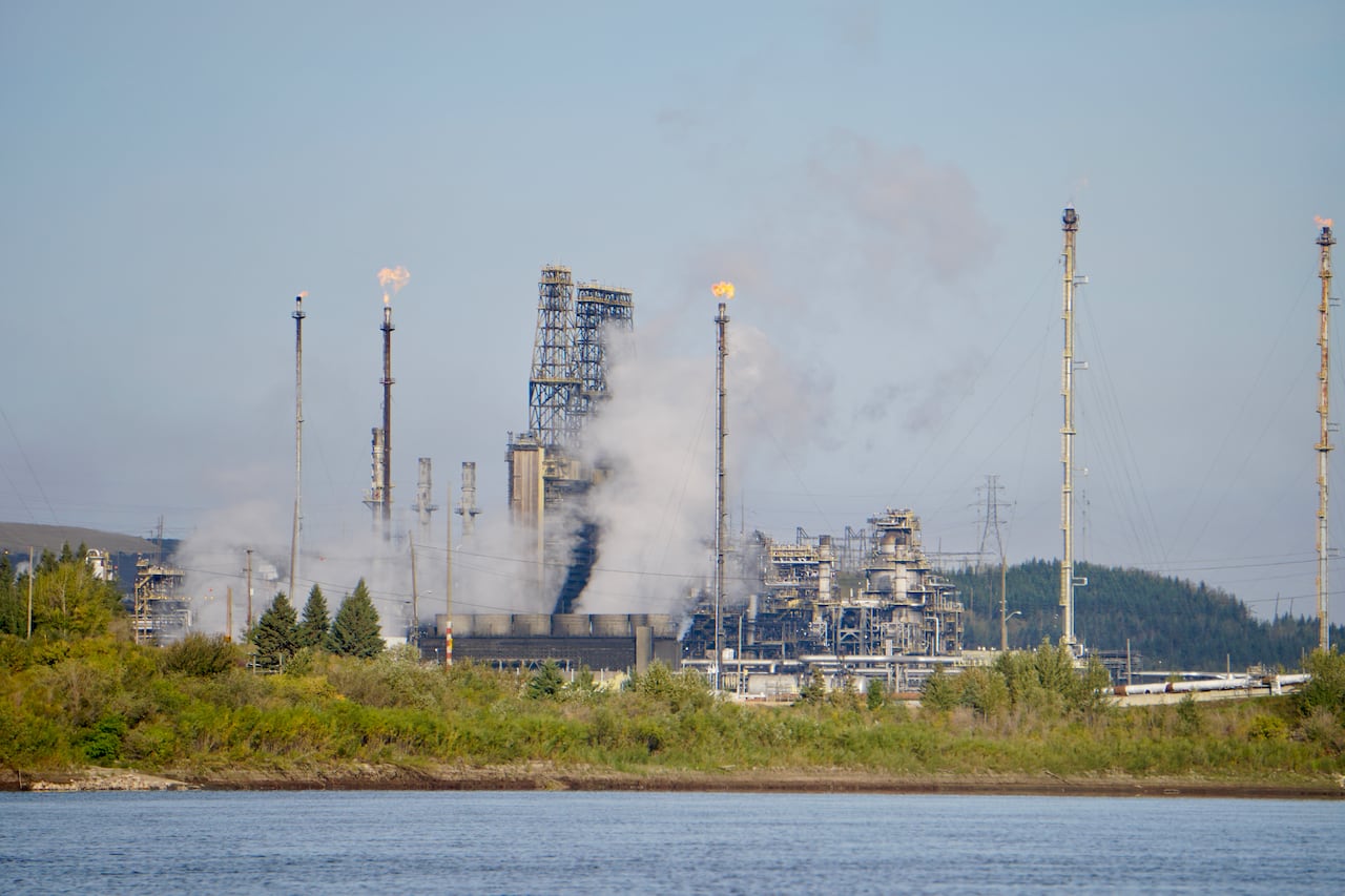 Flares are seen from smokestacks and industrial buildings, with a river in the foreground.