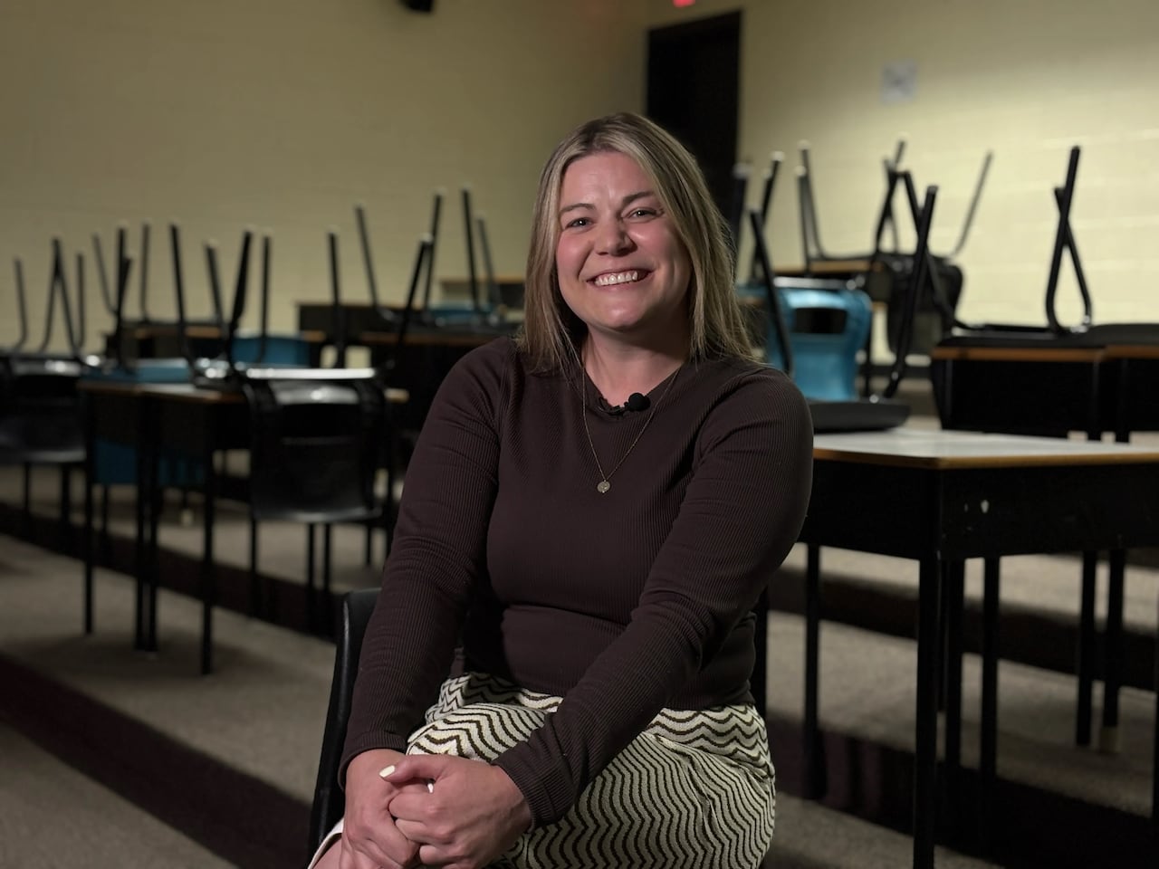 A smiling woman with long blonde hair wearing a brown sweater and patterned pants sits in a classroom, chairs overturned on desks behind her.