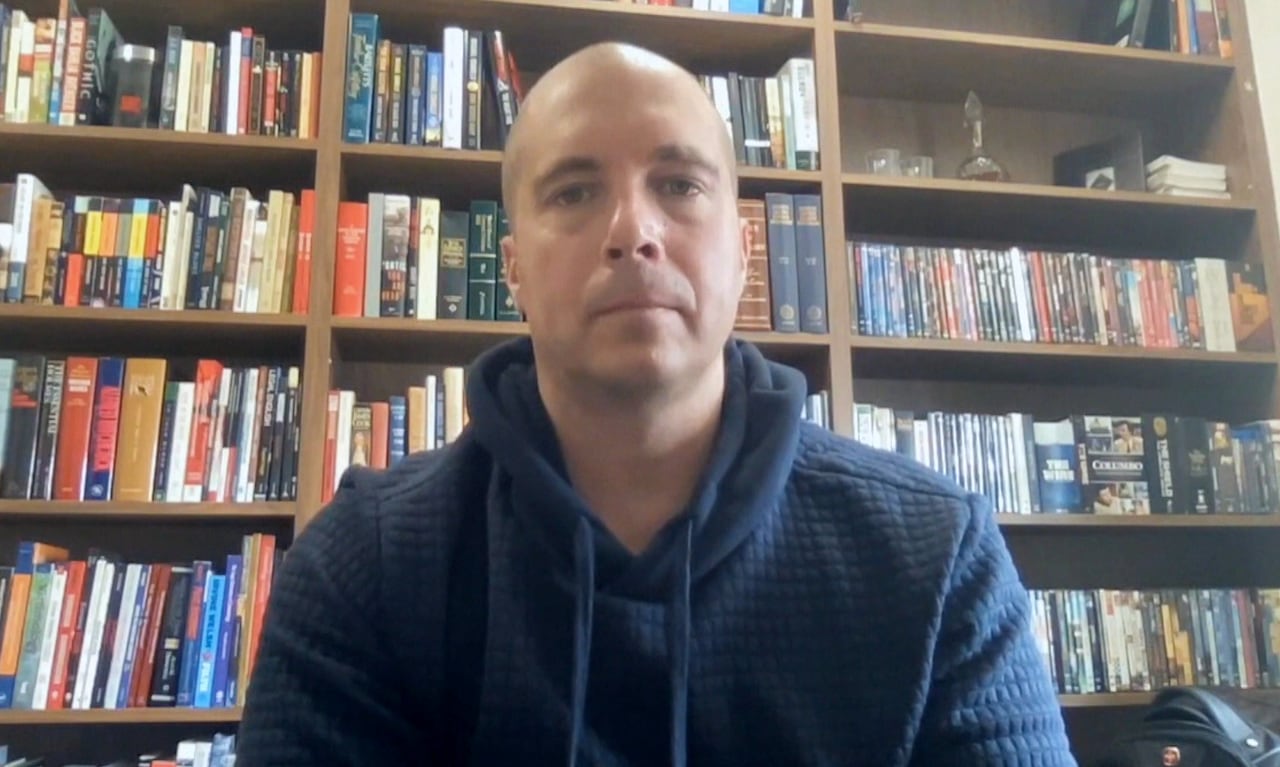 A bald man wearing a blue hooded sweater sits in front of a bookshelf.