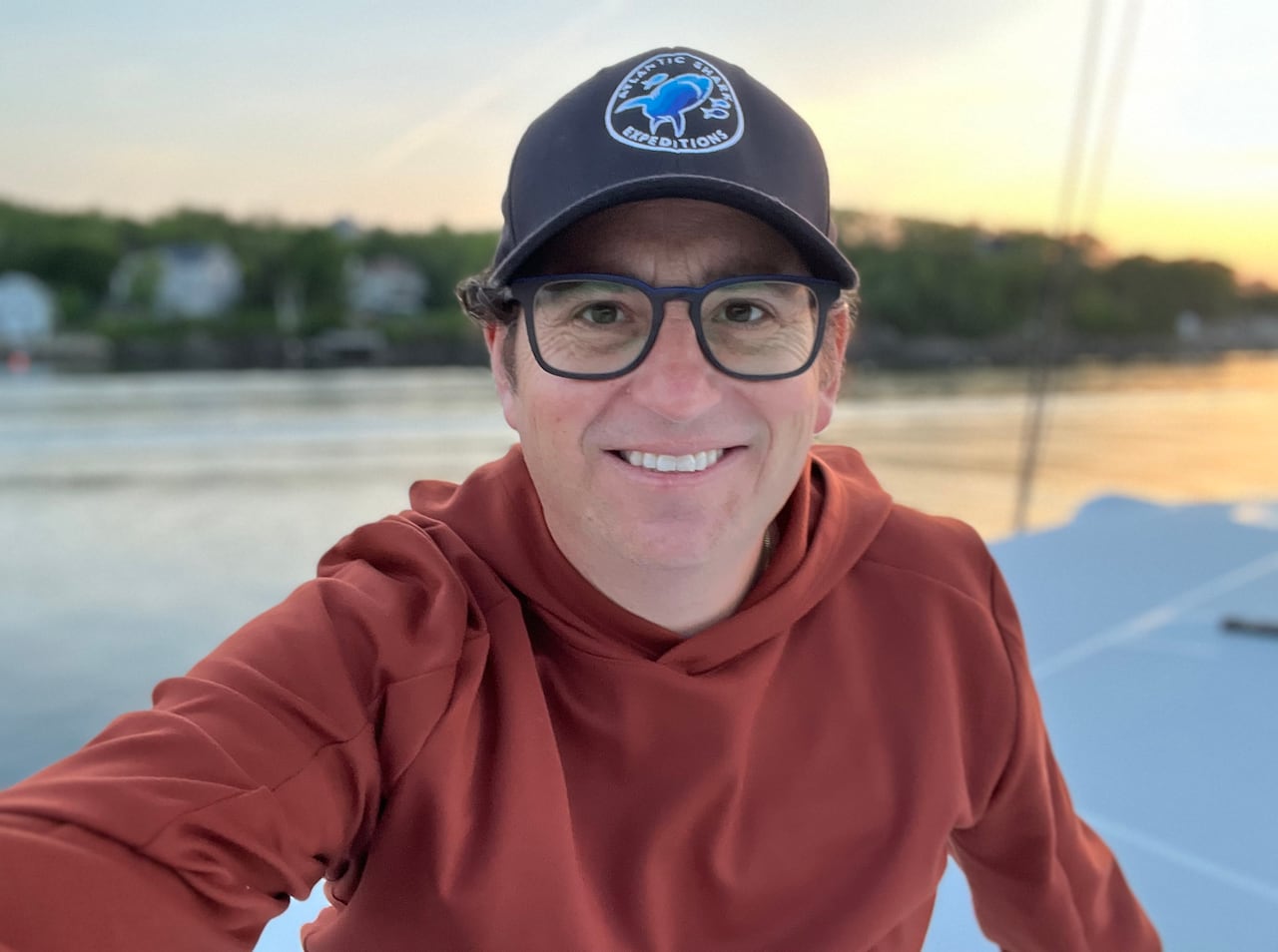 A smiling man with square glasses. He is wearing a baseball hat and an orange hoodie sitting on a boat.