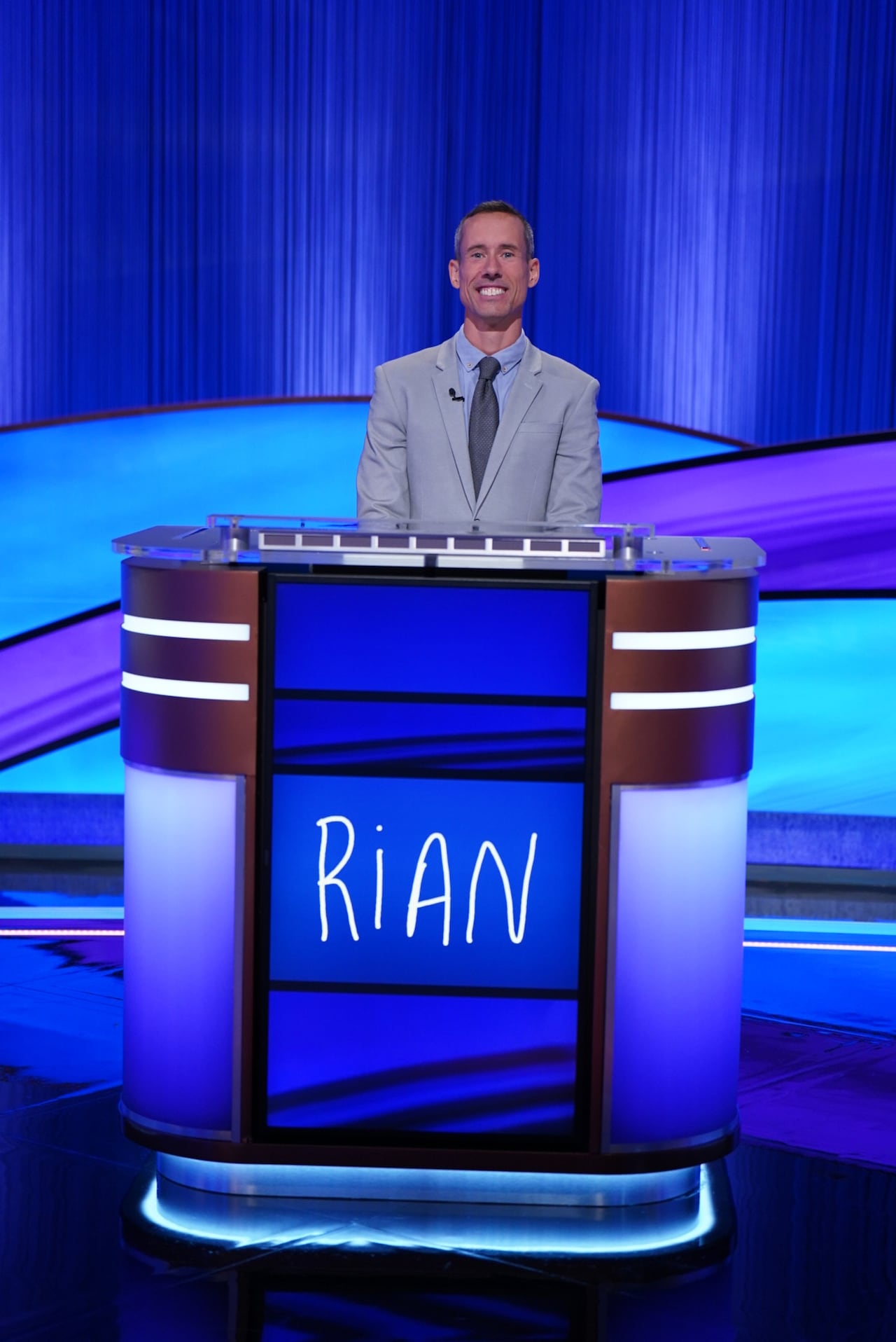 A man wearing a suit poses behind a podium with his name "Rian" spelled out on it.