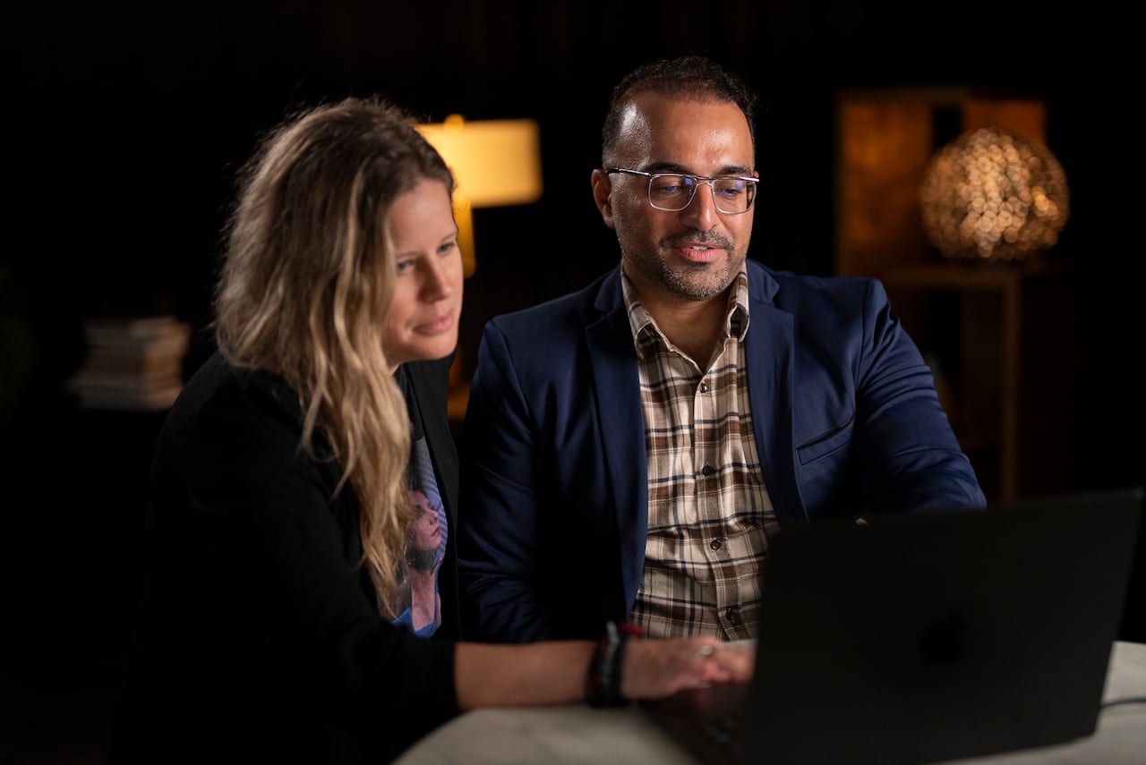 A woman and a man sit at a table and look at a black laptop.