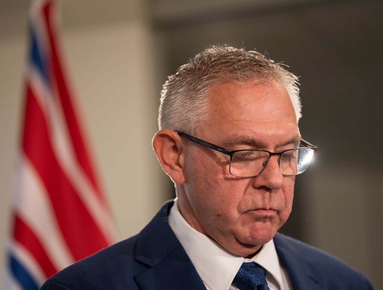 A white man wearing glasses is seen in front of a B.C. flag, looking down.