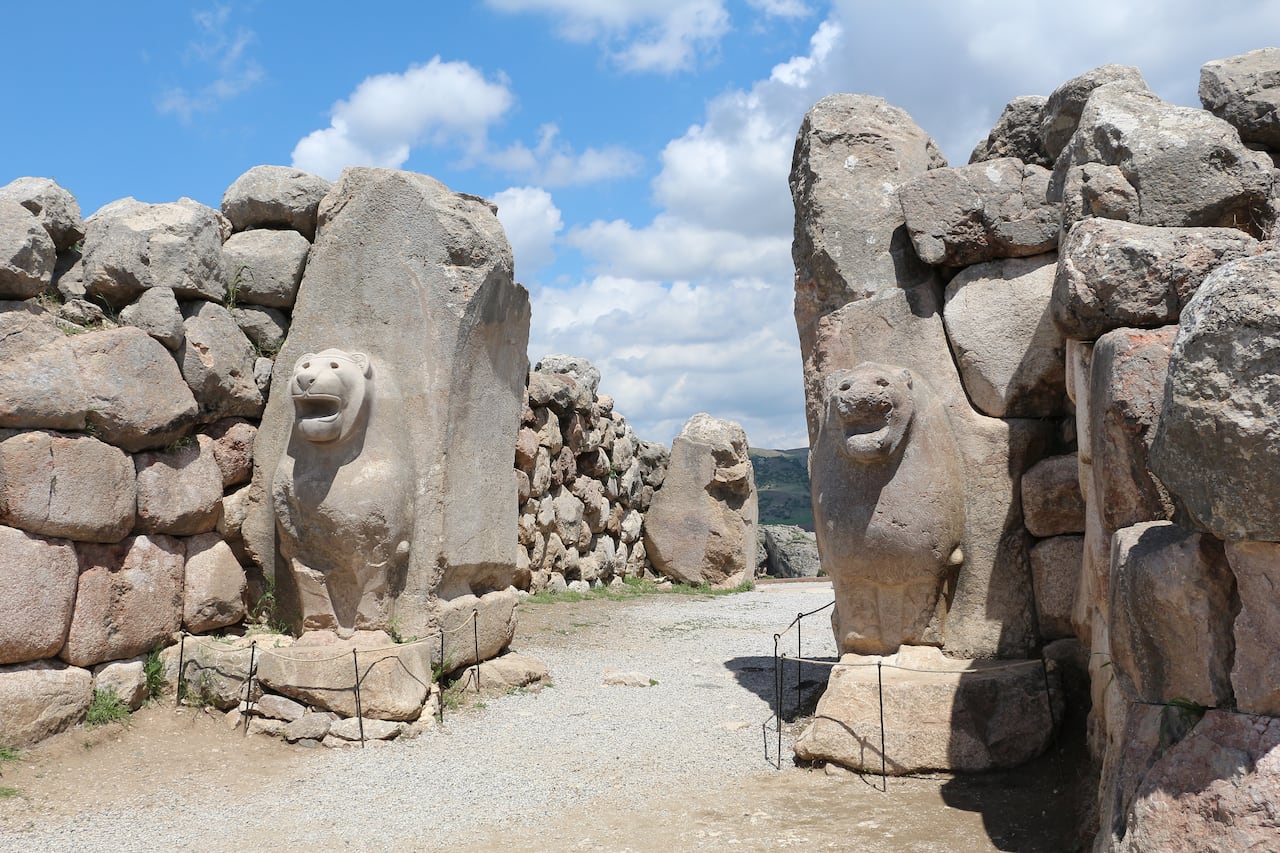 Two weathered stone lion sculptures flank the entrance to the ancient Lion Gate at Hattusa, with ruined walls and grassy hills in the background.