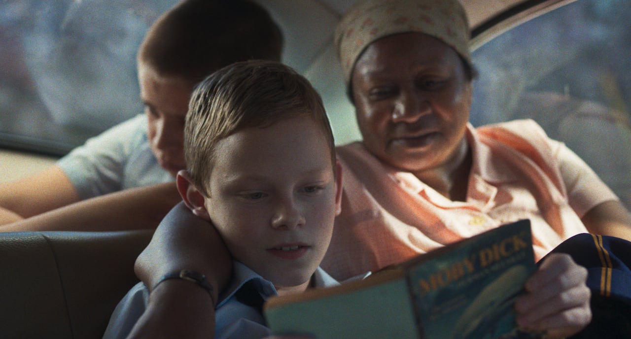A young boy, who is white reads Moby Dick in a car while an older woman, who is Black, puts her arm around his shoulder.