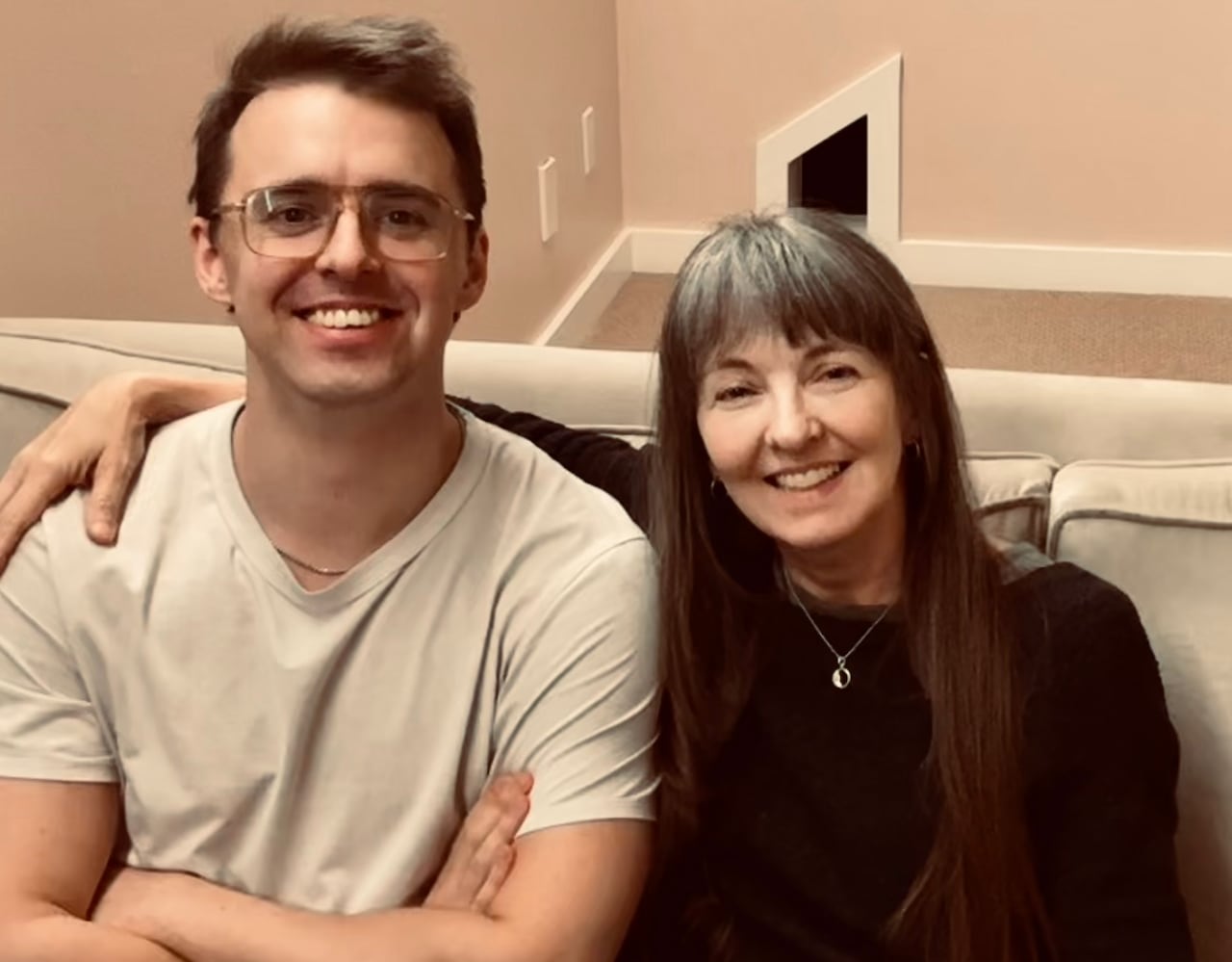 a young man with dark hair and glasses sits beside a middle aged woman with long dark hair on a couch, both smiling