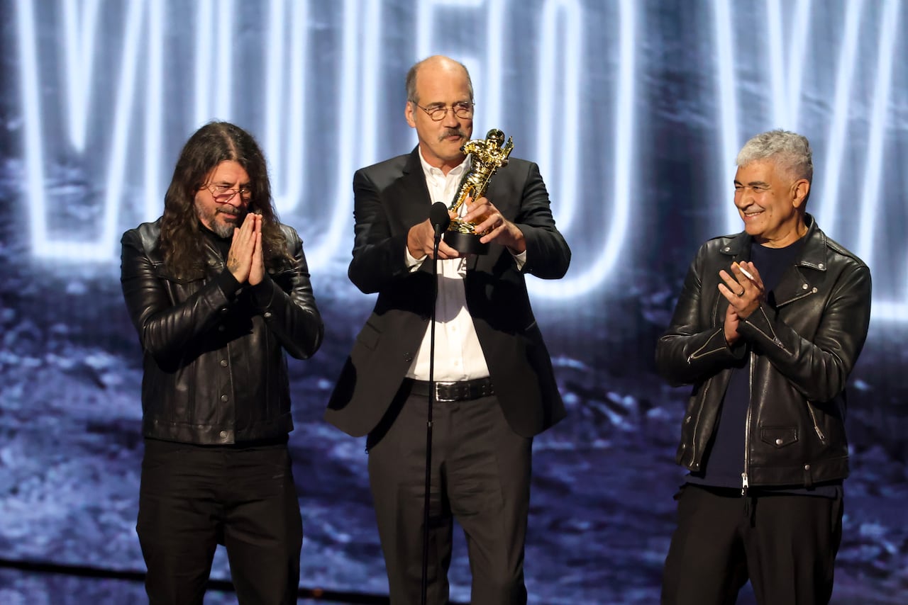 Three men on stage at a microphone. The one in the middle holds up a statuette.