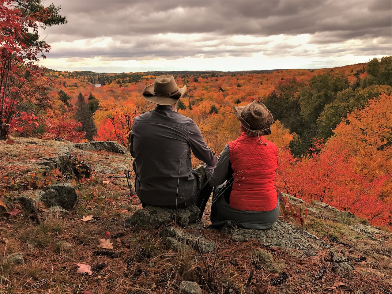 Two hikers wearing hats sit together on a rocky overlook above a forest filled with bright red, orange and yellow autumn leaves.