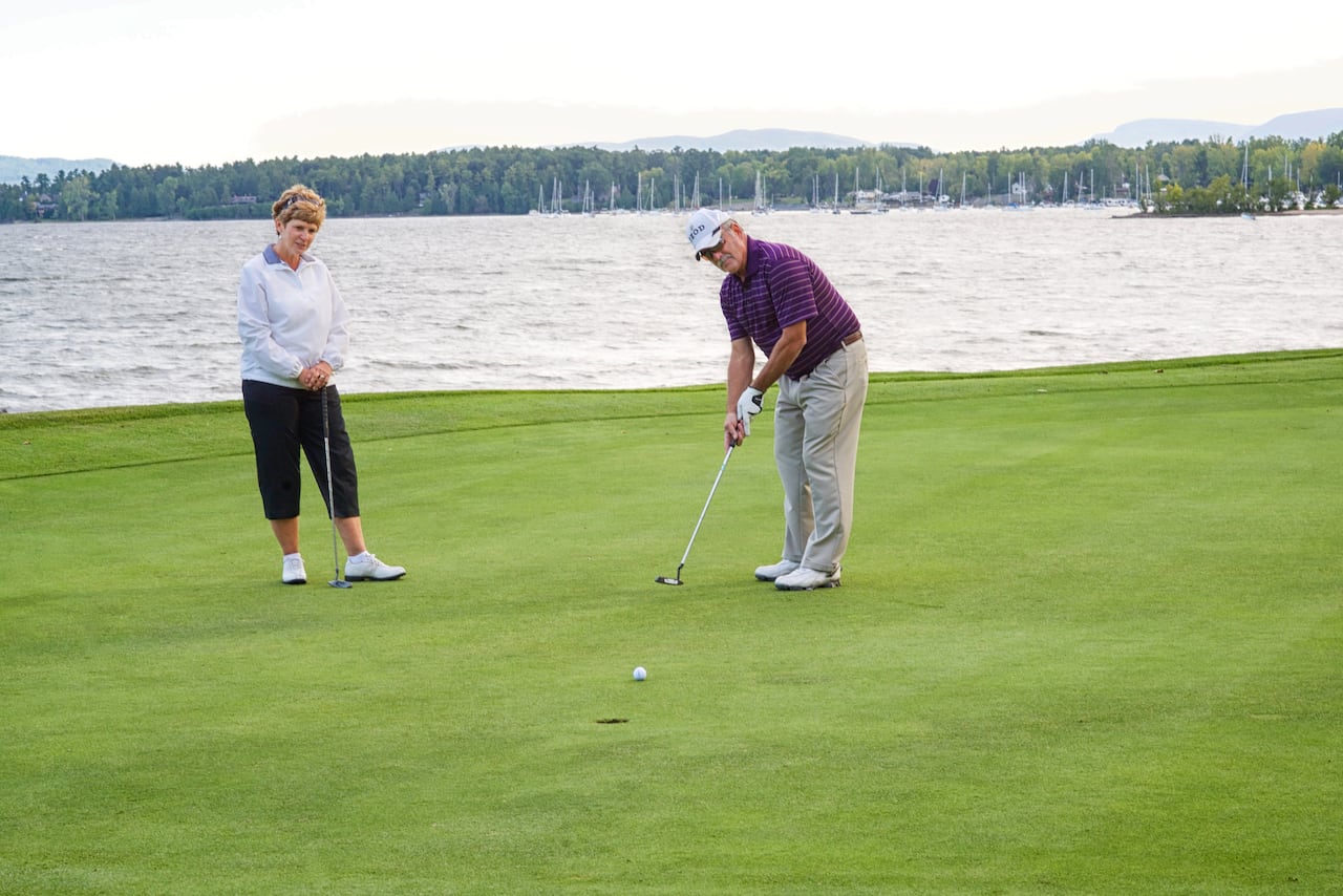 an older woman in black kapris and a grey sweater stands to the left watching an older man in a red polo shirt and white cap putt a golf ball towards a hole. in the background a body of water is visible behind the pair