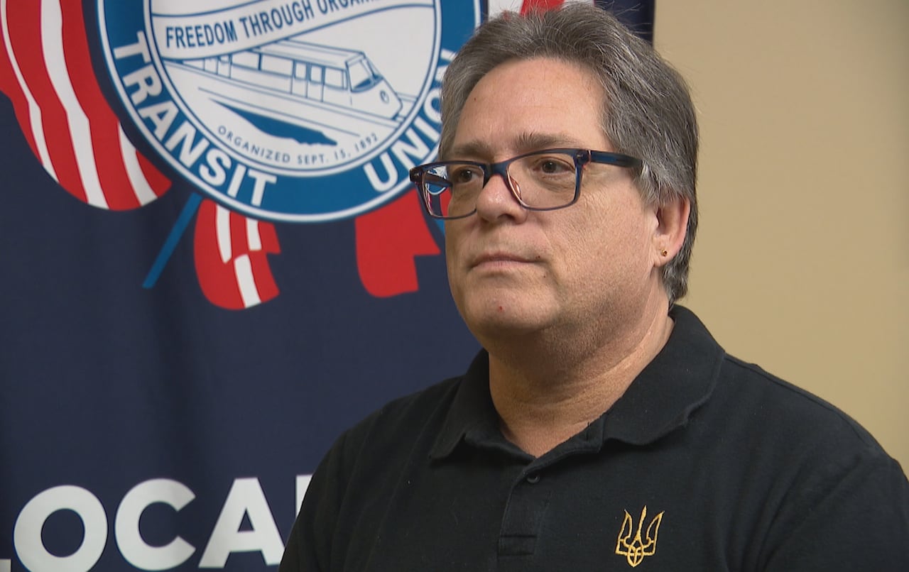 A man with grey hair wearing a black shirt and black, square glasses stands in front of a blue, red and white flag.