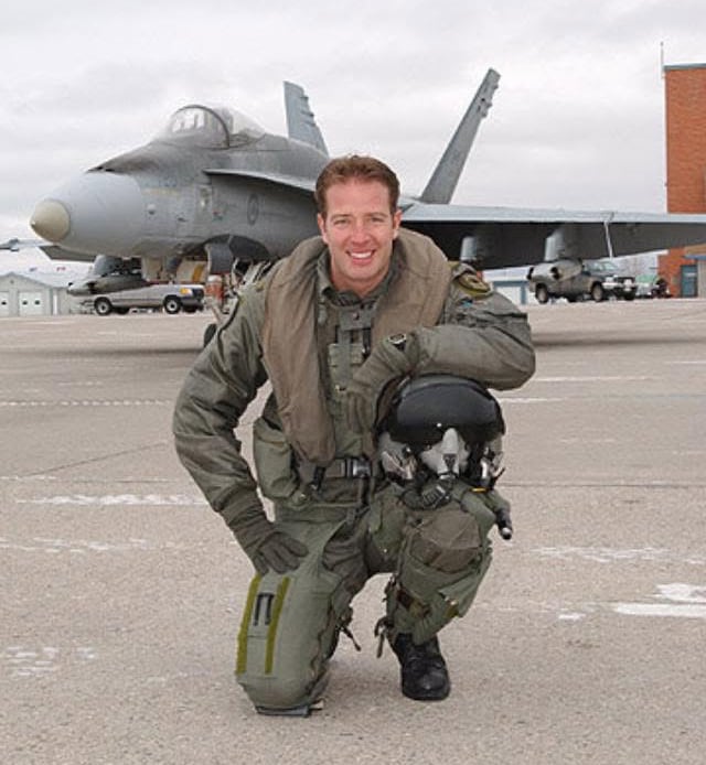 A fighter pilot kneels on the tarmac in front of a jet.