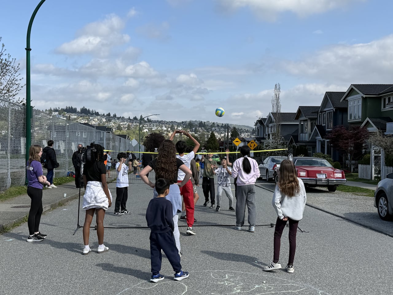A group of children play volleyball on a street closed to traffic. The sky is bright blue with some clouds.