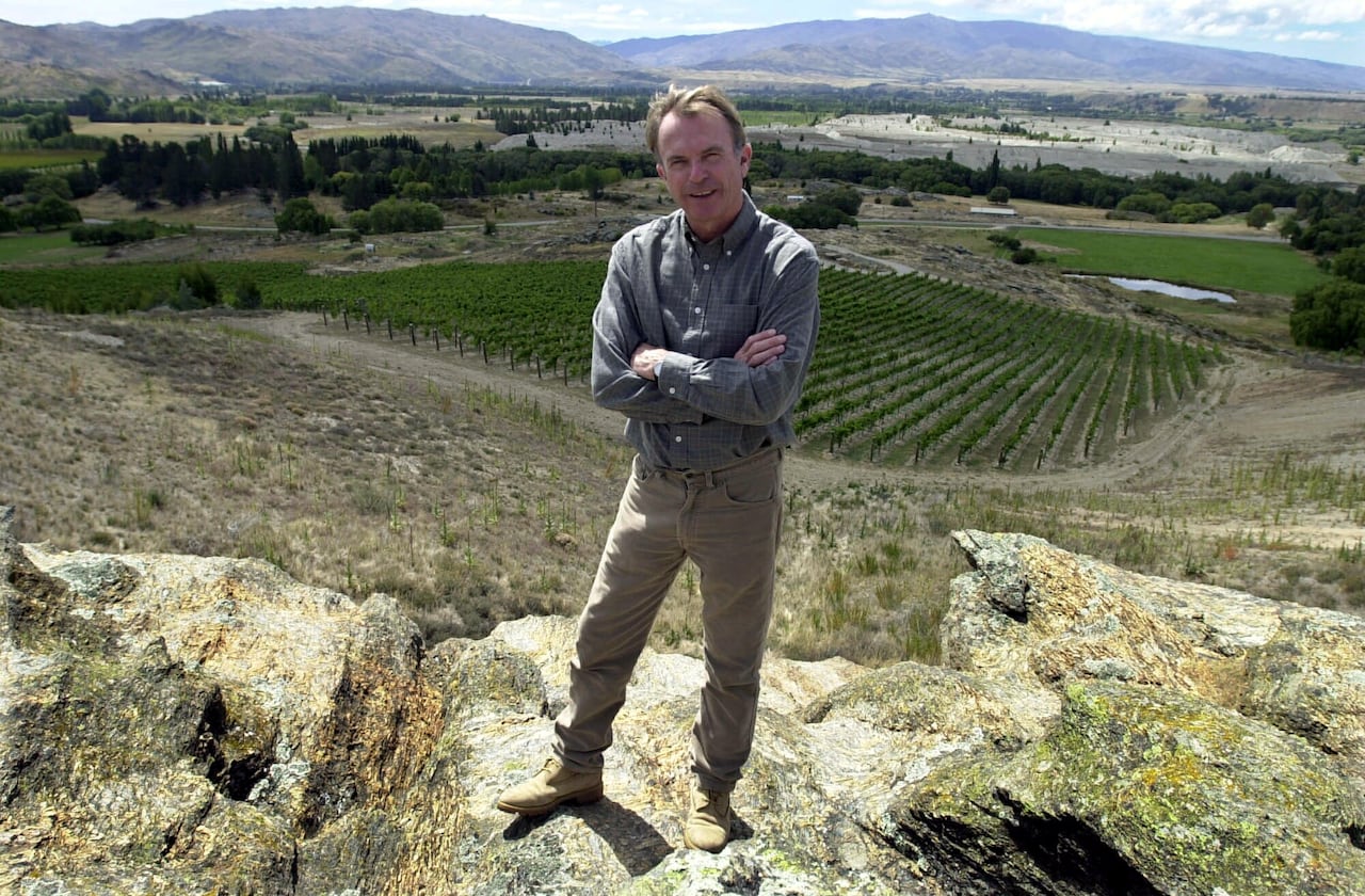 A cleanshaven man in a collared shirt and pants stands on a cresat above a wide expanse of rural land.