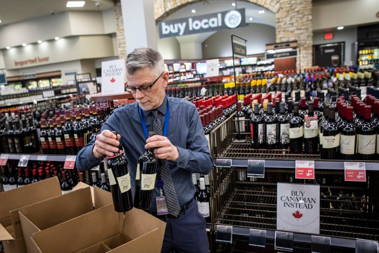 A man places bottles of wine in a box alongside a rack of bottles at a store. A 'Buy Canadian Instead' sign is visible.