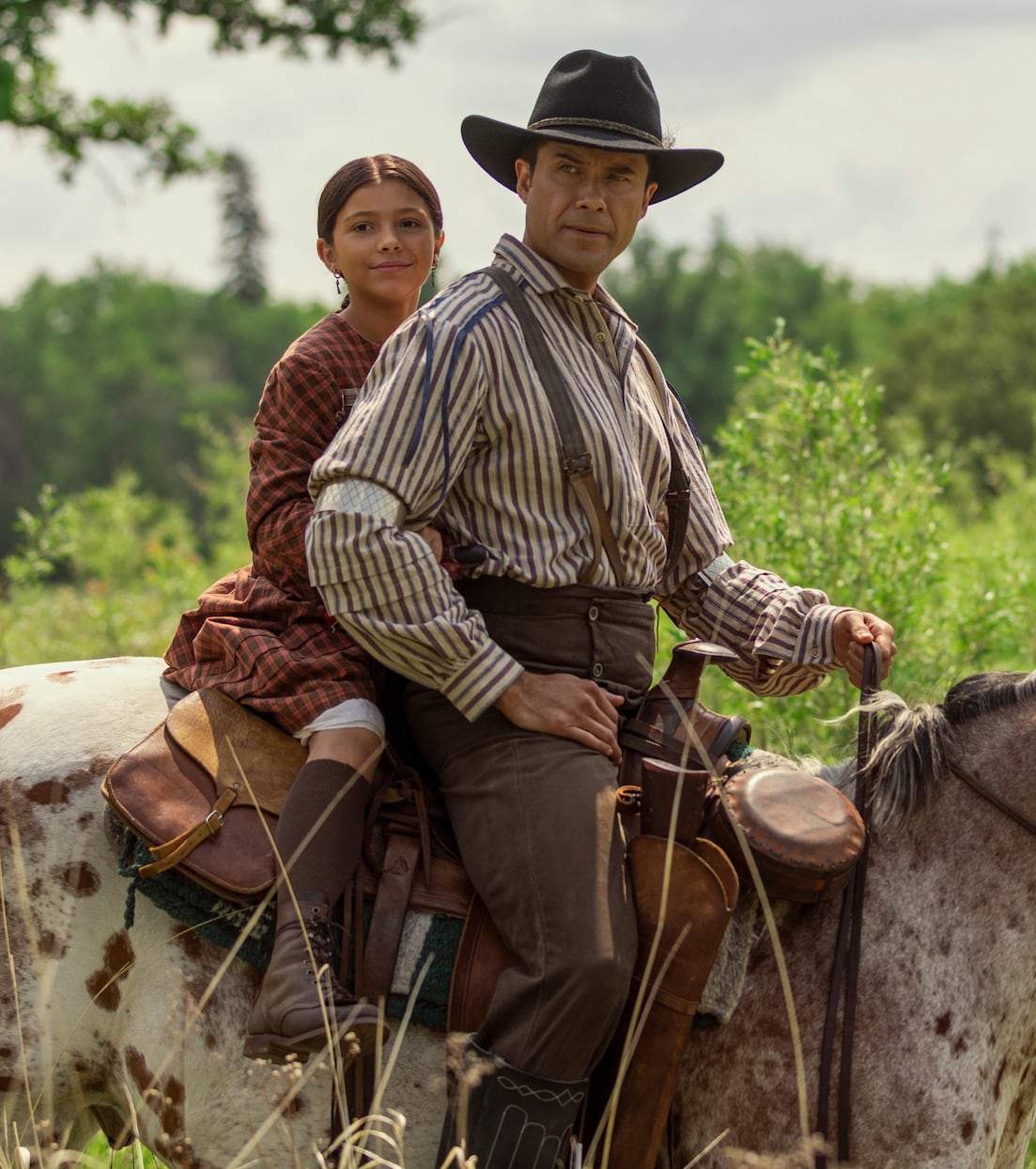 Indigenous man sitting on horse wearing a hat, with a young girl playing his daughter sitting behind him on the saddle.