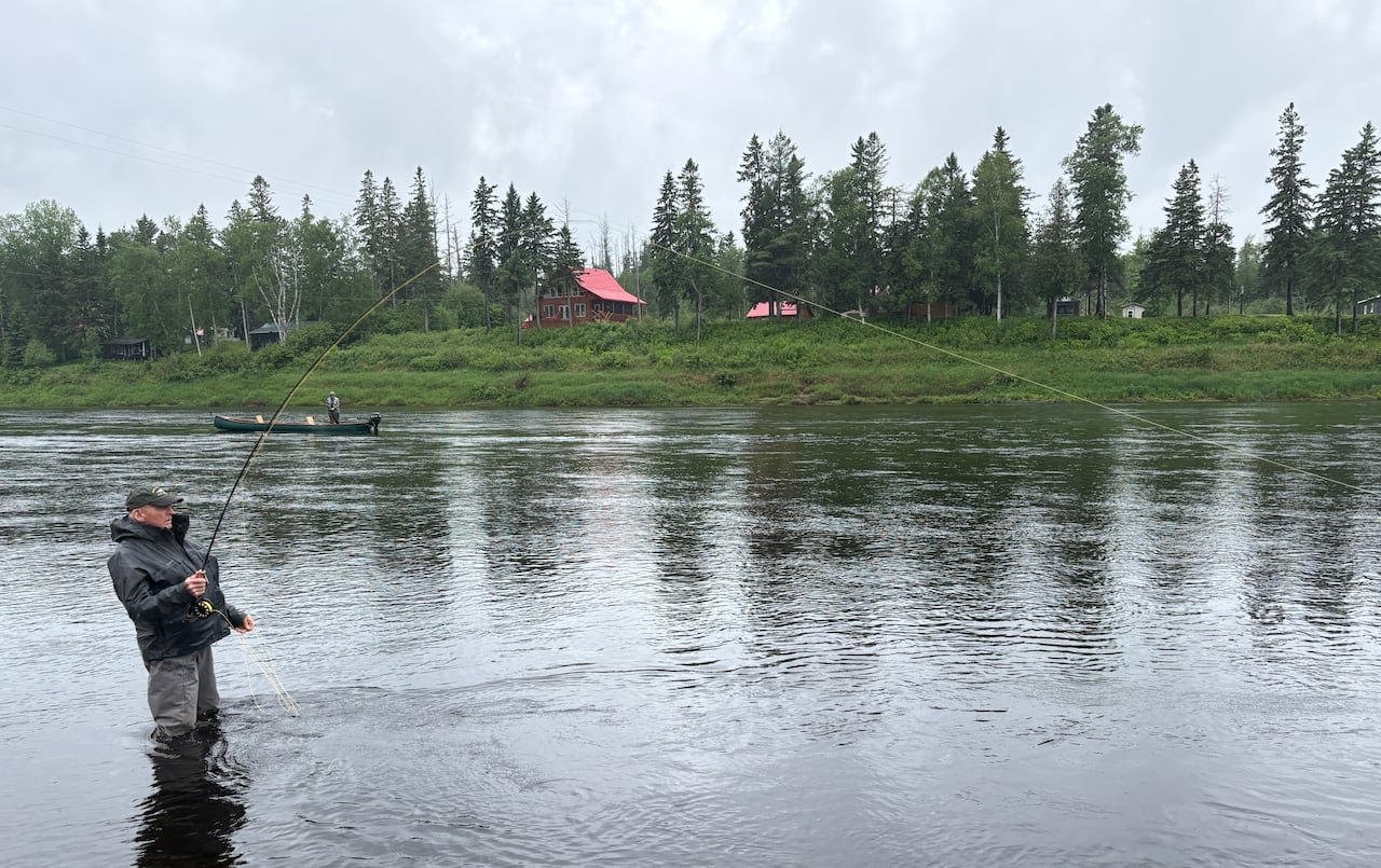 一名男子在河中钓鱼，背景是河岸和小屋。