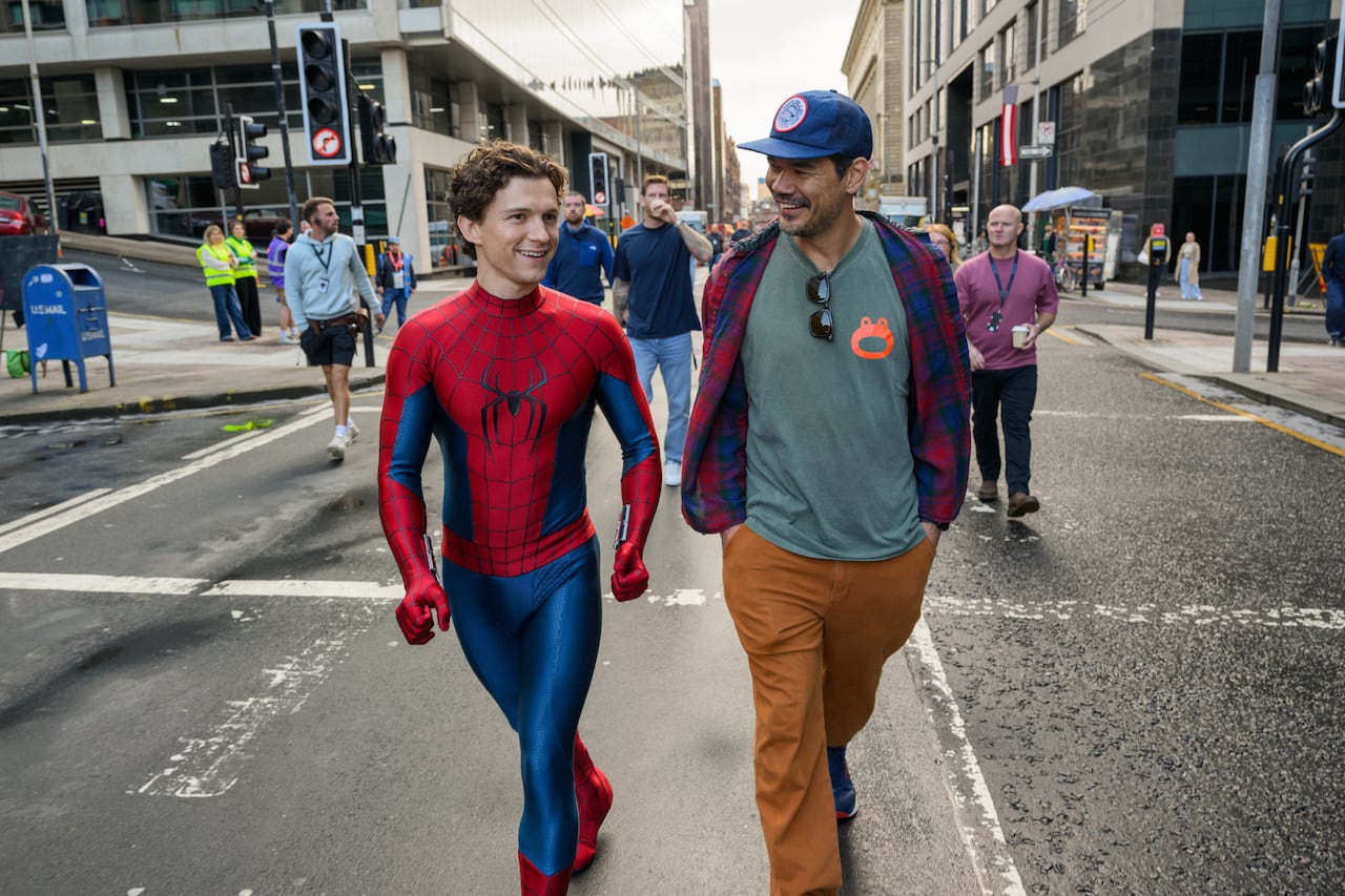 Actor Tom Holland and director Destin Daniel Cretton walk down a city street on the set of the new Spider-Man film.  Tom is in the Spider-Man costume with the mask off.