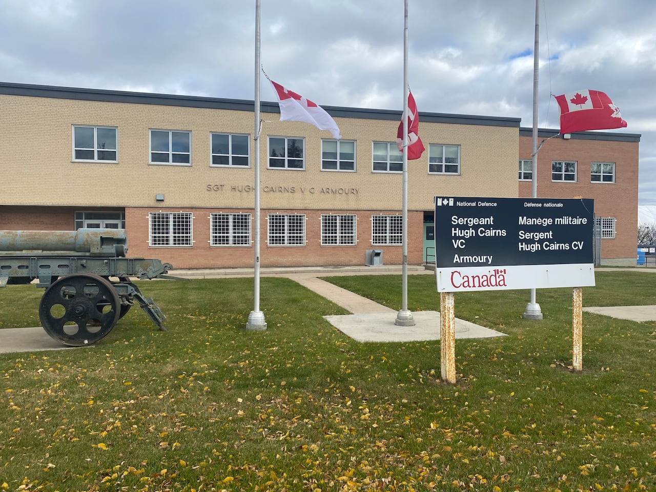 Canadian flags at half mast behind a sign that says "Sgt. Hugh Cairns VC Armoury."