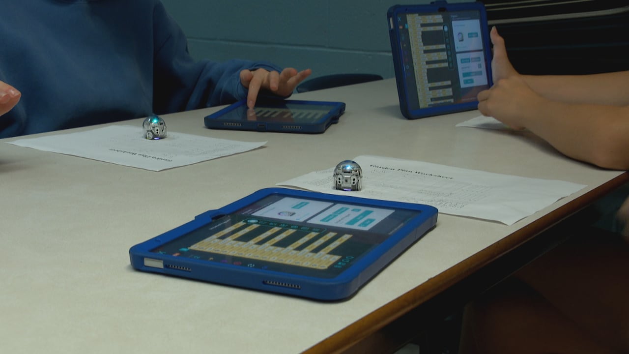 A close-up image shows the hands of students using iPads and sheets of paper to program tiny spherical robots.