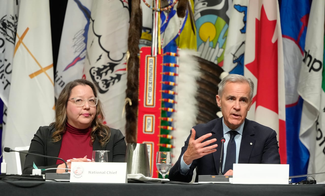 A woman with long brown hair, wearing glasses and a black blazer, sits beside a grey-haired man wearing a dark suit as he gestures while speaking into a microphone in front of many flags.