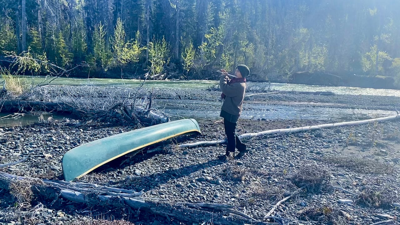 A man playing a flute beside a river and a canoe.