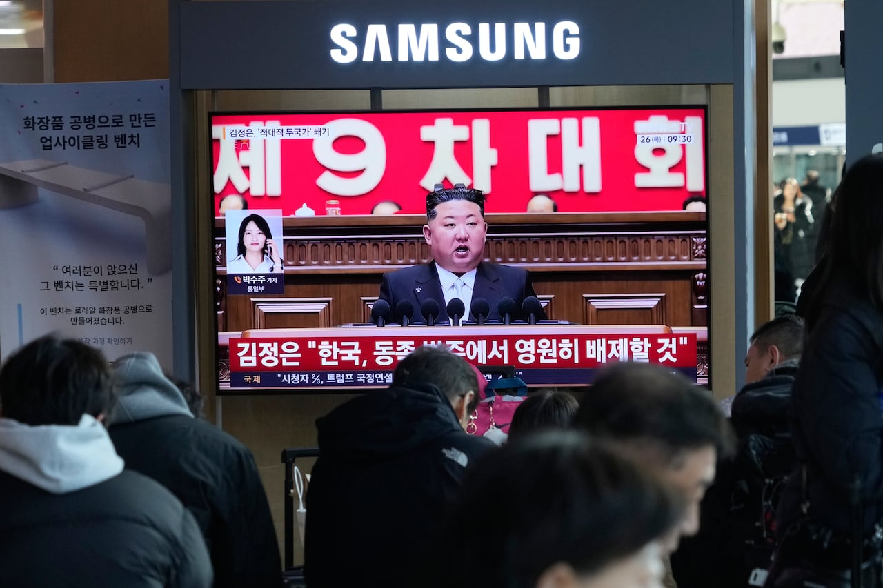 Man on a television screen speaks as people walk past.