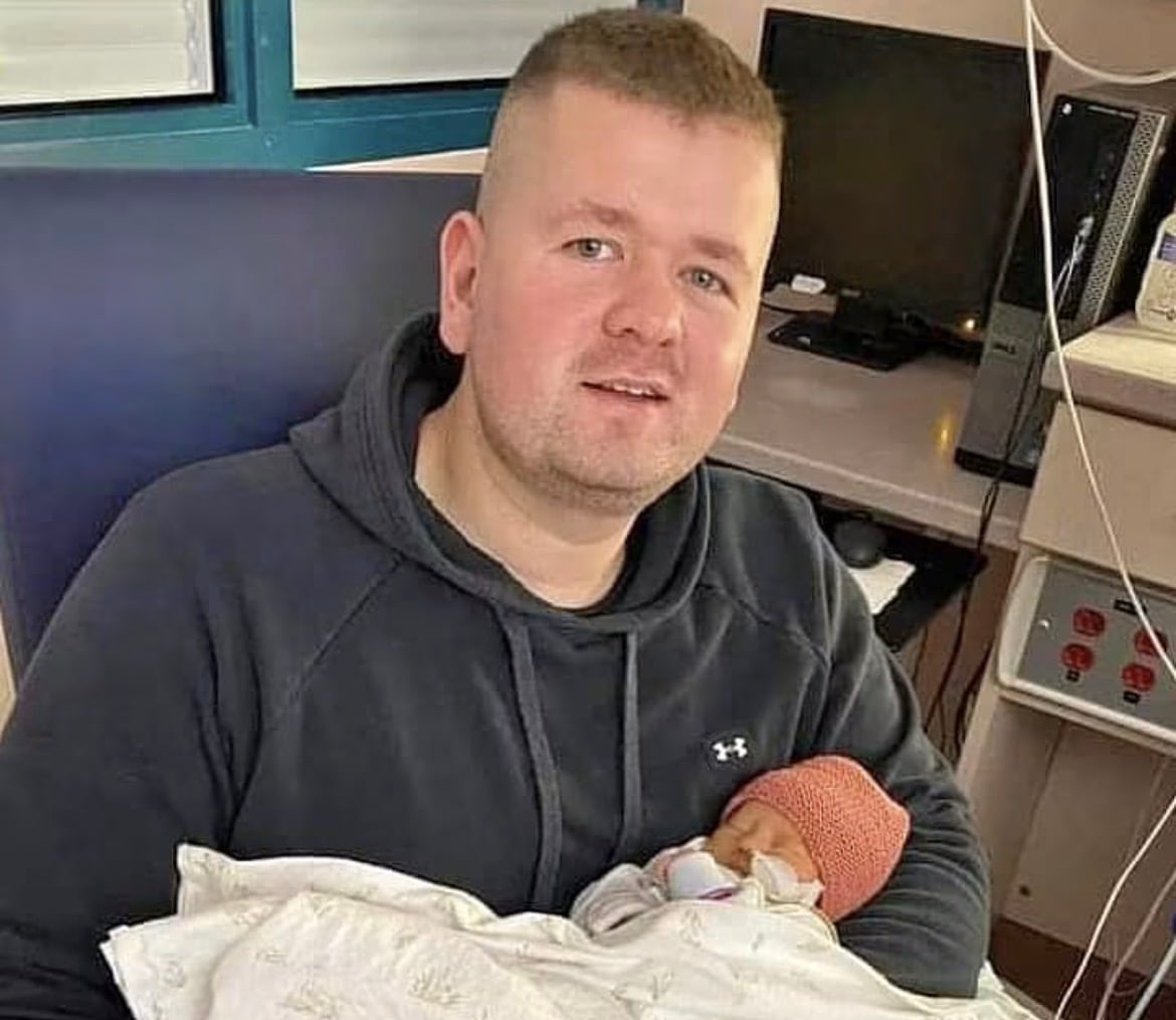 A smiling man holds his newborn baby in a hospital.