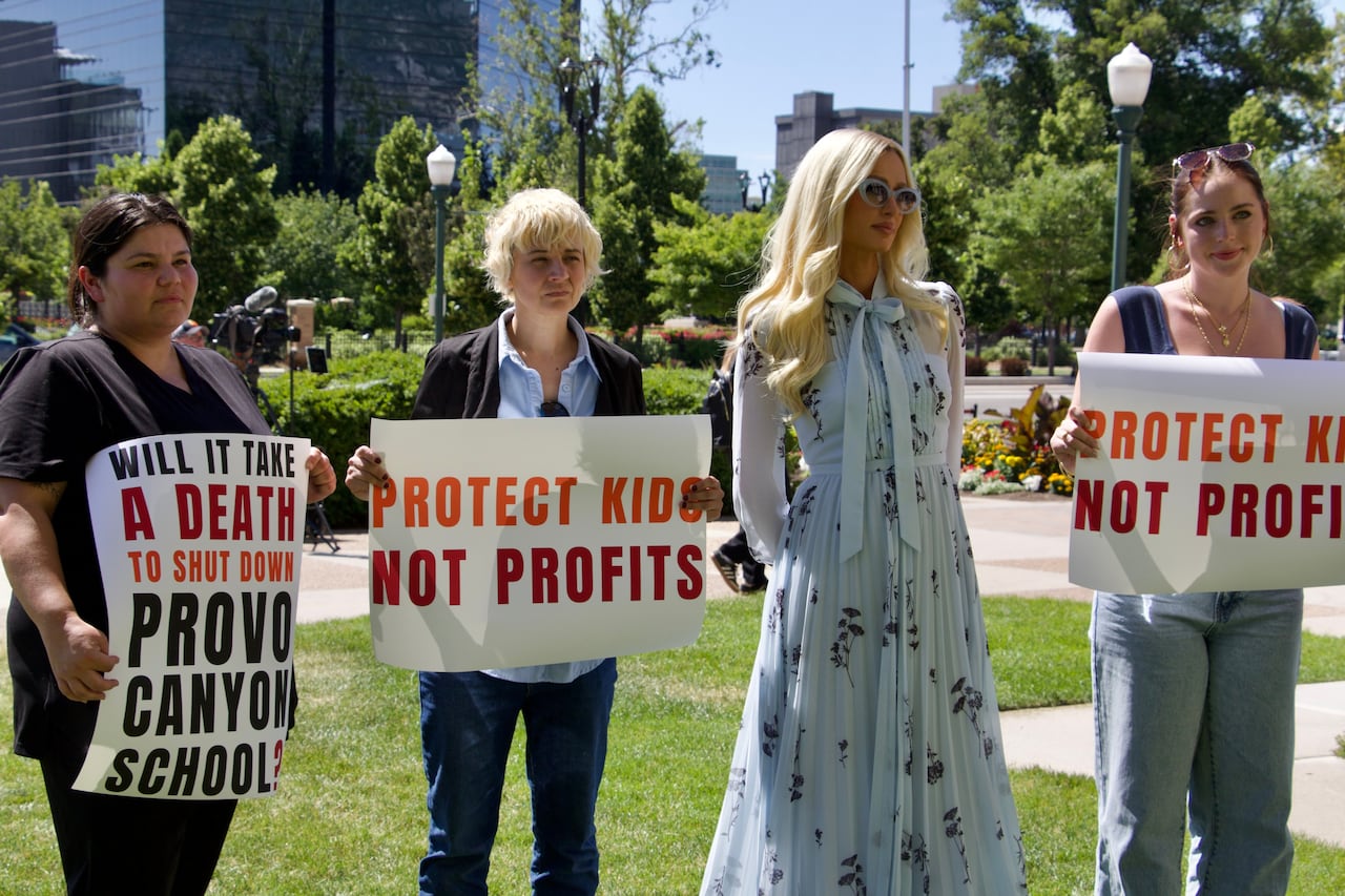 A woman in a long blue dress is flanked by younger people holding signs that read "protect kids not profits."