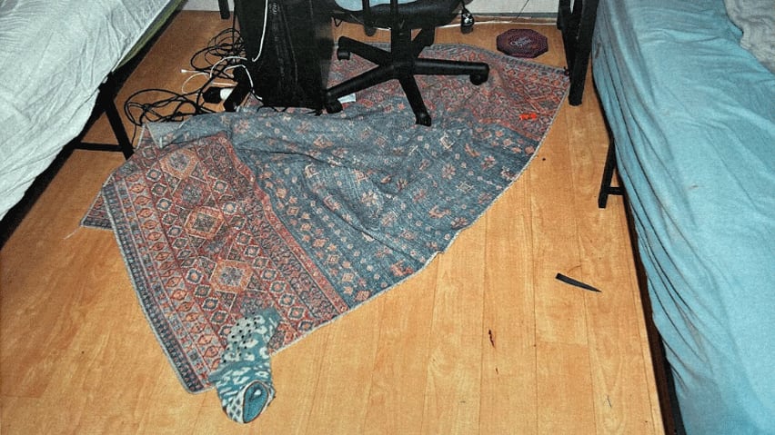 A close-up of the floor between two twin beds, showing a broken knife blade, a lone sock, a rumpled rug and a wrapped candy.