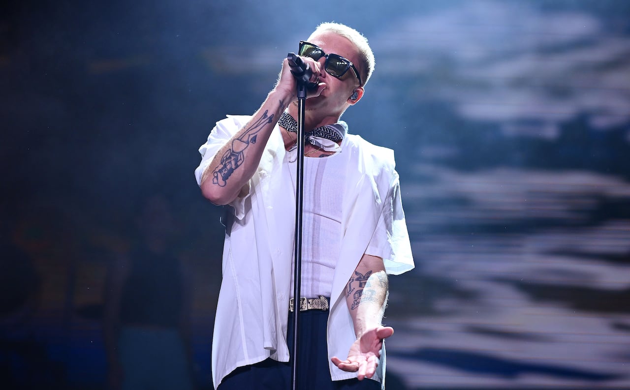 A singer in a white open button shirt, white tank top and black sunglasses