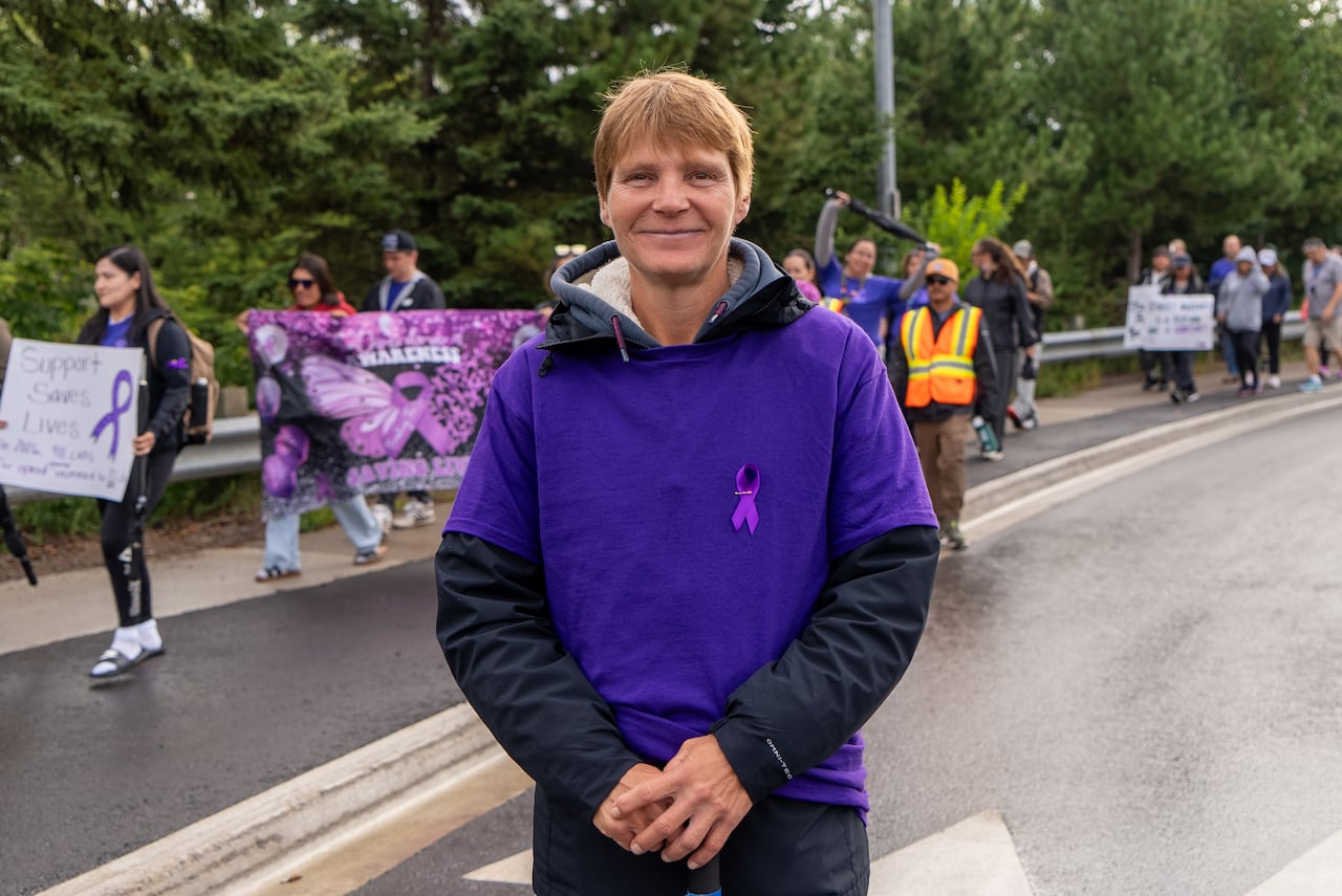 A person wearing purple is seen standing on a road. A group of people is seen walking behind them.