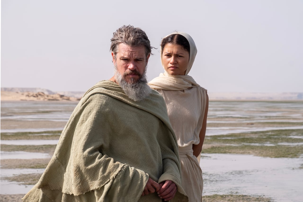 A man and woman in simple robes stand on a beach.