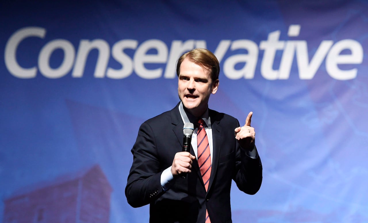 Man in suit holding microphone and finger up in front of Conservative backdrop