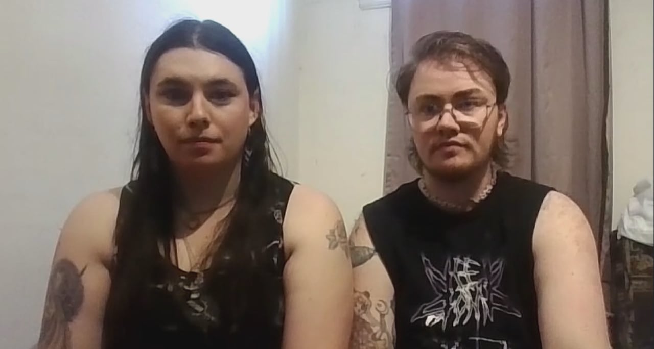 Two people sit in a white room with a window blind hanging behind them.